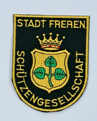 Das Bild zeigt das Abzeichen der Schützengesellschaft der Stadt Freren. Das Emblem im Wappen ist das der Stadt Freren, ist auf dunklem Hintergrund gestaltet und umfasst eine goldene Krone über einem Wappen mit einem dreiblättrigen Kleeblatt. Der Schriftzug „Stadt Freren Schützengesellschaft“ umrah