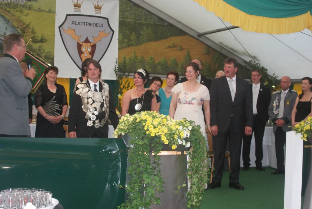 Feierliche Szene beim Schützenfest in Freren: Mitglieder der Schützengesellschaft stehen zusammen, während eine Ansprache gehalten wird. Im Hintergrund ist das Vereinswappen zu sehen, umrahmt von festlicher Dekoration und Blumen.