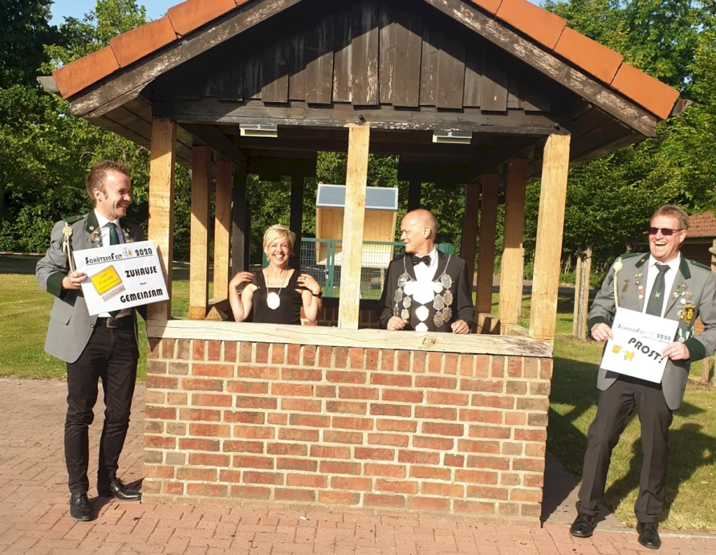 **Alternativbeschreibung:** 

Vier Mitglieder der Schützengesellschaft Freren stehen an einem festlich dekorierten Kiosk und halten Schilder mit den Aufschriften „Zuhause gemeinsam“ und „Prost!“ in den Händen. Die Szene strahlt Freude und Gemeinschaftsgeist während des Schützenfestes 2020 aus.