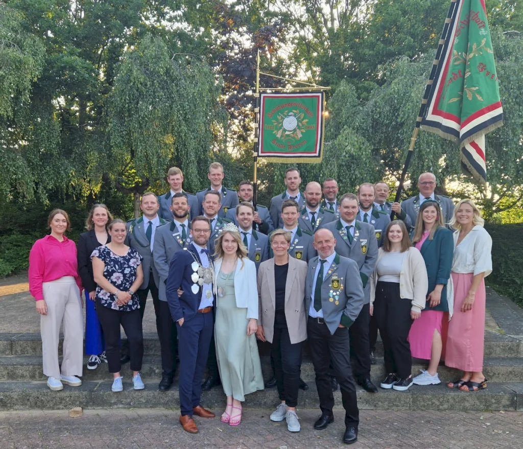 Gruppenfoto der Schützengesellschaft Freren beim Schützenfest. Die Mitglieder in festlicher Kleidung posieren vor der Fahne der Gesellschaft, umgeben von grüner Natur. Ein Symbol für Gemeinschaft und Tradition in Freren.